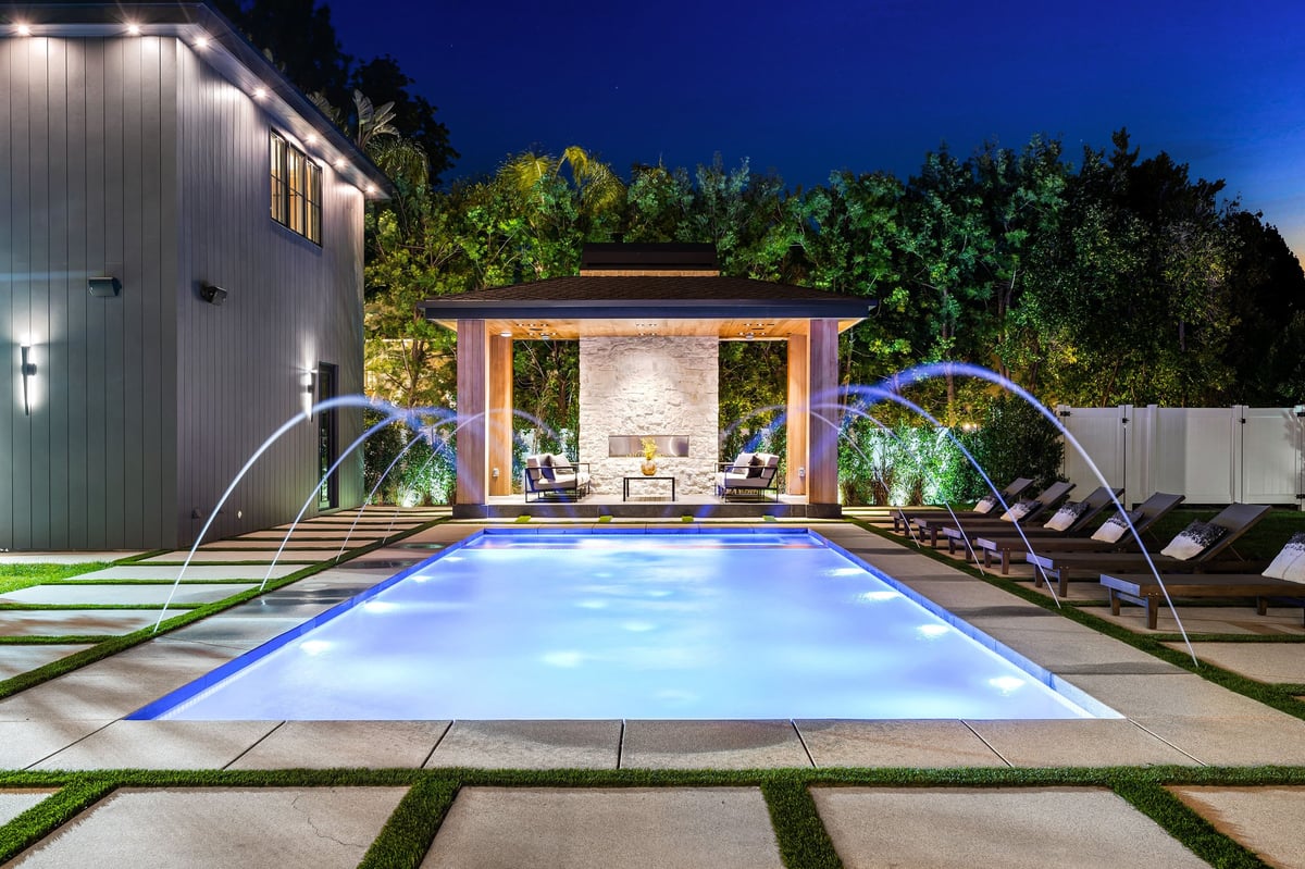 Luxurious modern backyard with illuminated swimming pool, lounge chairs, and a covered seating area at night