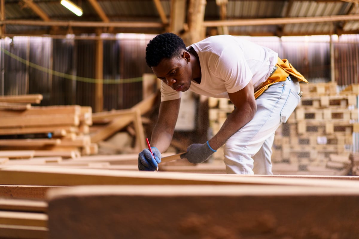 An African carpenter in a woodworking workshop carefully inspecting a sawn wooden plank with focus and precision