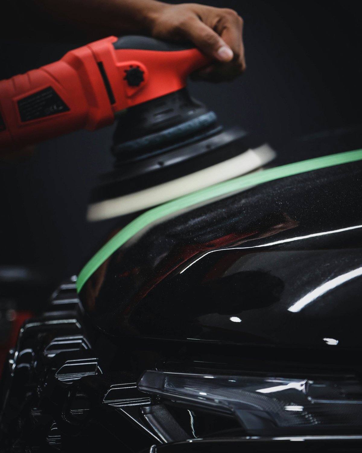 Person using red orbital sander on black car hood with green masking tape visible