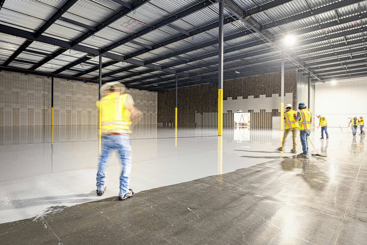Construction workers in yellow safety vests cleaning a large industrial warehouse floor with water spray