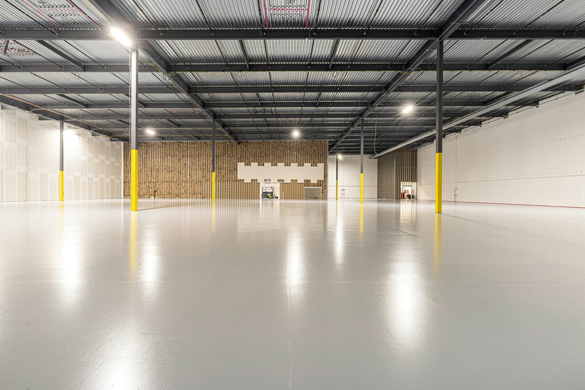 Spacious industrial warehouse interior with polished concrete floor, metal ceiling structure, yellow support columns, and bright overhead lighting.
